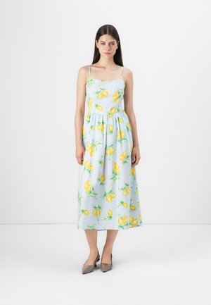 Maxi dress in light blue fabric with lemon pattern, thin straps, gathered waist, and a flowy silhouette. Worn with gray heels.