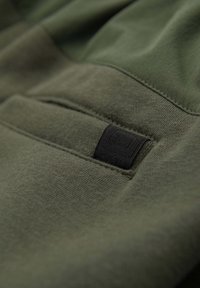 Gros plan sur un tissu vert olive avec une étiquette tissée noire cousue près d'une couture, mettant en valeur la texture et les détails de la couture.