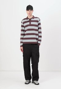 Polo de mangas longas à risca em bordeaux e branco, com colarinho e lista de botões, combinado com calças cargo pretas e ténis cinzentos.