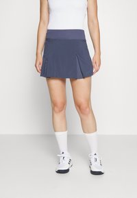 Falda deportiva azul marino con una cinturilla ancha, diseño plisado, tejido que absorbe la humedad, combinada con calcetas blancas hasta la rodilla y zapatillas deportivas.