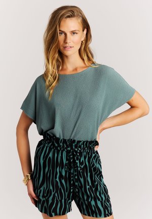 Femme portant un haut ample à manches courtes bleu sarcelle, rentré dans un short taille haute bleu sarcelle et noir à imprimé animal, posant avec la main sur la hanche.