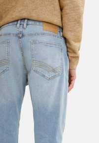 Jeans din denim de culoare albastru deschis, cu talie medie, având două buzunare la spate, decorate cu cusături aurii și o etichetă de brand din piele bej.