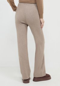 Person, die beigen, weit geschnittenen Strickhosen, ein braunes langärmliges Oberteil und dunkellange Plattformschuhe trägt, steht auf einem weißen Boden.