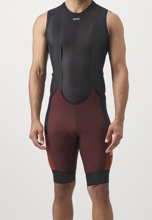 Mężczyzna sportowiec w bezrękawnikowych czarno-bordowych spodenkach kolarskich na szelkach, stojący na jednolitym tle.