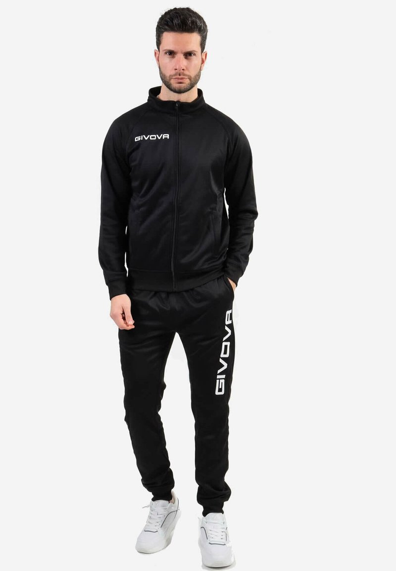 Tuta sportiva nera realizzata in tessuto morbido; giacca con zip e colletto alto e logo, pantaloni coordinati con logo lungo il lato. Sneakers bianche.