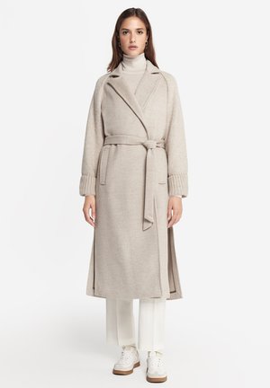 Ung kvinde iført beige uldfrakke med bælte, lys rullekrave, hvide bukser og hvide sneakers, stående mod en hvid baggrund.