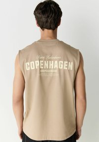 T-shirt sans manches beige avec un grand imprimé "COPENHAGUE" au dos, accompagné d'un texte plus petit en haut et en bas dans une police contrastée.