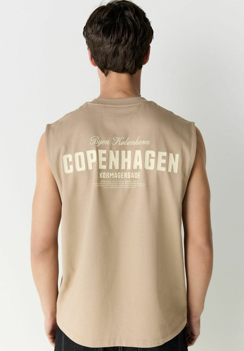 T-shirt sans manches beige avec un grand imprimé "COPENHAGUE" au dos, accompagné d'un texte plus petit en haut et en bas dans une police contrastée.