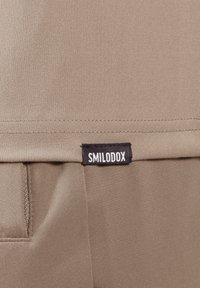 Primer plano de una tela color beige con una etiqueta negra de "SMILODOX". El material es suave, con un acabado mate y textura mínima.