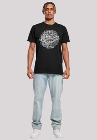 Black cotton t-shirt featuring a white graphic design in a circular layout. Paired with light denim jeans and white sneakers.