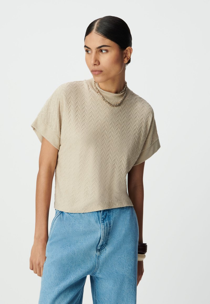 Sandałowy, krótkowany sweter z teksturowanym wzorem w rybie łuski, z krótkimi rękawami i wysokim dekoltem, w połączeniu z jasnoniebieskimi dżinsami.