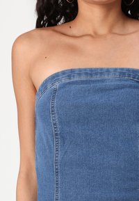 Denim top na tubu v srednji modri barvi. Gladka tekstura z dobro prileganje. Surov rob na vrhu in vertikalni šivi. Brez naramnic.
