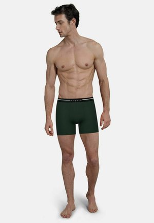 Muskulöser Mann steht barfuß in dunkelgrünen Boxershorts mit "bugatti"-Logo am Bund und blickt nach links vor einfarbigem Hintergrund.