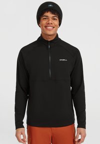 Zwarte half-zip pullover gemaakt van een gladde, stretchstof. Beschikt over lange mouwen en een wit logo op de borst. G gedragen met een oranje onderstuk.