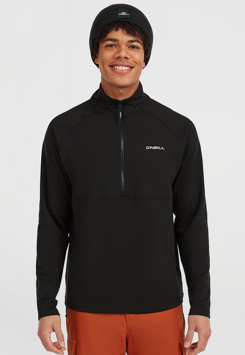 Zwarte half-zip pullover gemaakt van een gladde, stretchstof. Beschikt over lange mouwen en een wit logo op de borst. G gedragen met een oranje onderstuk.