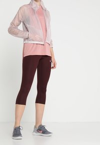 Lichtgewicht, doorschijnende roze jas over een roze geperforeerd top, gecombineerd met aansluitende burdgundy leggings en grijze sportschoenen.