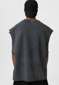 Grijze mouwloze shirt met een losse pasvorm, met een vervaagde textuur en brede armsgaten. Gecombineerd met een zwarte pet.