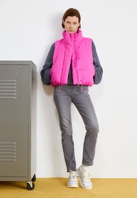 Gilet imbottito rosa con alto colletto, bottoni a pressione e orlo con cordino, abbinato a jeans grigi e sneakers chiare, su uno sfondo neutro.