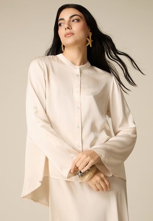 Mujer con cabello largo y oscuro, con blusa de seda color crema con botones, falda a juego, pendientes de estrella de mar y varias pulseras, mirando hacia un lado.