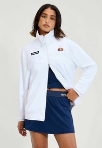 White zip-up jacket with embroidered logo on chest, paired with a navy skirt. Features a soft texture and a relaxed fit design.