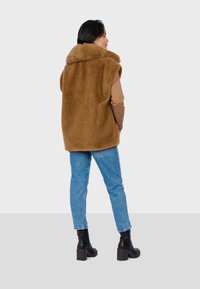 Gilet in finta pelliccia marrone con un ampio colletto, indossato sopra una maglia beige a maniche lunghe. Abbinato a jeans in denim blu chiaro e stivali neri con tacco alla caviglia.