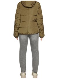 Olijfgroene gewatteerde jas met capuchon, gestikte uitstraling en elastische manchetten, gecombineerd met lichtgrijze spijkerbroek en beige schoeisel.