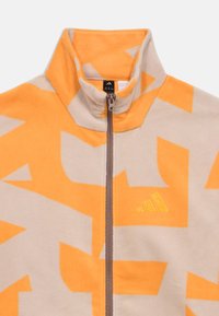 Ζακέτα φλις με μπεζ βάση και μεγάλα πορτοκαλί γεωμετρικά σχέδια. Διαθέτει ψηλό γιακά και μπροστινό φερμουάρ. Λογότυπο Adidas σε κίτρινο χρώμα.