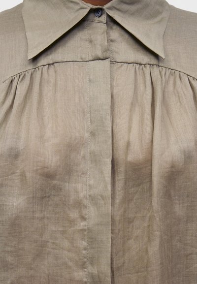 Μπεζ λινή μπλούζα με μυτερό γιακά, κρυφή πατιλέτα με κουμπιά και ρυτιδωμένες λεπτομέρειες υφάσματος κάτω από το φιτίλι.