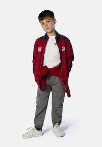 Chaqueta con cremallera roja y azul marino con emblemas, camiseta blanca, pantalones cargo grises con bolsillos y zapatillas blancas con cordones. Tejido texturizado.