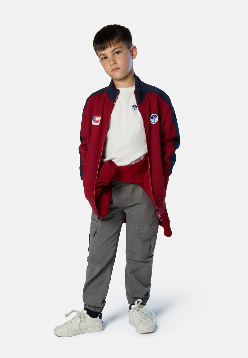 Chaqueta con cremallera roja y azul marino con emblemas, camiseta blanca, pantalones cargo grises con bolsillos y zapatillas blancas con cordones. Tejido texturizado.