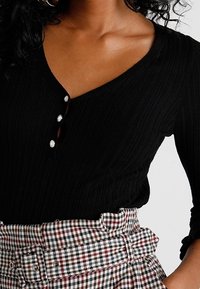 Czarny prążkowany top z długim rękawem, dekoltem w serek i trzema białymi guzikami. Połączony z plisowaną spódnicą w kratę z paskiem, w kontrastujących kolorach czerwonym, czarnym i białym.