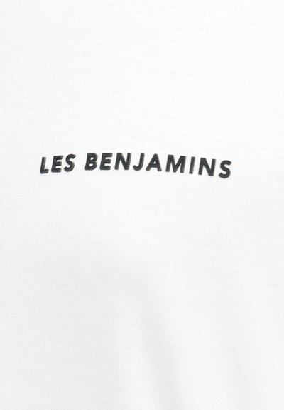 Λευκό ύφασμα με μαύρο κεφαλαίο κείμενο «LES BENJAMINS» τοποθετημένο στο κέντρο της επιφάνειας.