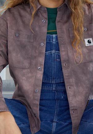 Henkilö, jolla on siniset farkkuhaalarit, vihreä paita alla ja ruskea napitettava Carhartt-takki.