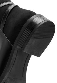 Botín negro que combina cuero liso y gamuza suave, con un tacón de goma texturizada. El tacón tiene ranuras horizontales para mayor agarre.