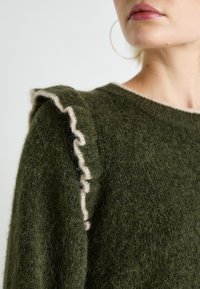 Gros plan sur une personne portant un pull duveteux vert olive avec des bords volantés et une légère bordure, une grosse boucle d'oreille visible.