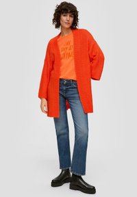 Oranger Strickcardigan mit weiten Ärmeln, kombiniert mit einem orangen Grafikshirt und blauen Jeans. Schwarze Knöchelstiefel runden das Outfit ab.