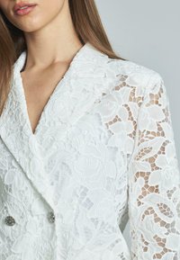 Blazer bianco in pizzo con motivi floreali, bottoni doppi e maniche in pizzo trasparente, che mostra una texture e dettagli di design intricati.
