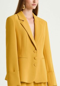 Blazer jaune sur mesure en tissu lisse. Il présente un col net, trois boutons sur le devant, et de larges poignets. Détails simples avec des poches.