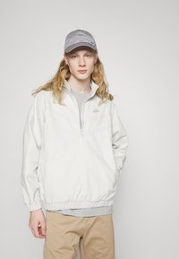 Hvid zip-up jakke med elastiske manchettter og kant, med en grå Nike-logo. Båret over en grå skjorte, parret med beige bukser og en grå kasket.