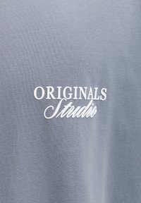 Γκρι βαμβακερό t-shirt με λευκή κεντημένη επιγρα φή «ORIGINALS Studio» στο μπροστινό μέρος, με απαλό υφή και casual σχεδιασμό.