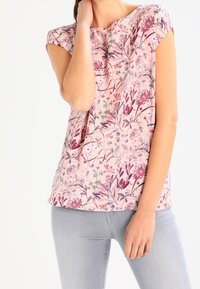 Blouse à manches courtes florale en rose doux avec des motifs violets et verts. Tissu léger, coupe décontractée, décolleté rond, poignets retroussés.
