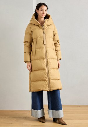 Long manteau rembourré tan, doté d'une capuche, d'une fermeture éclair à l'avant et de deux poches latérales. Porté avec un pantalon en denim à larges revers et des bottes à talons.