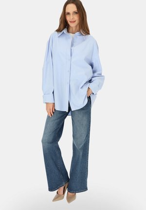 Kobieta stoi ubrana w jasnoniebieską oversize koszulę zapinaną na guziki, jeansy z szerokimi nogawkami i beżowe szpilki, pozując z jedną ręką w kieszeni.