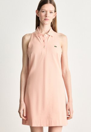 Vrouw in een mouwloze lichtroze polo jurk met kraag en knopen, staand tegen een effen lichte achtergrond.