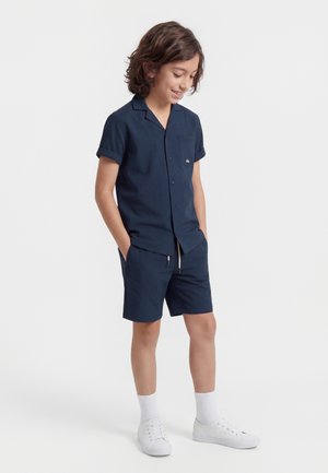 Chlapec so stredne dlhými vlasmi, oblečený v námornícky modrej krátkorukávovej košeli na gombíky a zodpovedajúcich šortkách, nosí biele ponožky a tenisky, ruky má v kapsách.