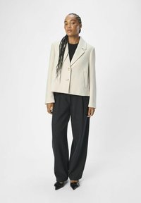 Chaqueta recortada de un beige claro con cuello, cierre de un solo botón y bolsillo lateral, combinada con pantalones negros holgados y zapatos negros puntiagudos.