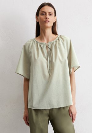 Vrouw met lang bruin haar die een losse lichtgroene korte mouwen blouse met een strik aan de hals en olijfgroene broek draagt, staand tegen een witte muur.