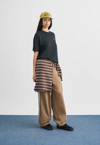 Schwarzes kurzärmliges Hemd, braun gestreifter Wickelrock, beige weite Hosen, schwarze Schuhe, gelbe Kappe mit Logo. Einfacher und lässiger Look.