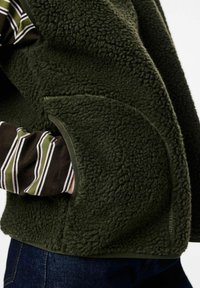 Persona valkā tumši zaļu flīsa vestu ar roku kabatā virs svītrainas garām piedurknēm krekliem un ziliem džinsiem.