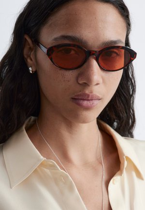 Femme aux cheveux bruns ondulés portant des lunettes de soleil ovales à monture écaille avec des verres rouges, des boucles d'oreilles argentées en forme de créole, une chemise beige à col et un mince collier argenté.